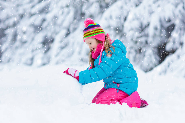 Kid making snowman in snowy winter park. Children play in snow. Little girl in colorful jacket and hat building snow man in winter garden after snowfall. Outdoor fun on cold winter day. - Zdjęcie, obraz