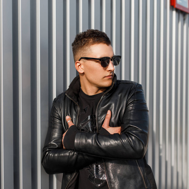 Beau jeune homme à la mode avec des lunettes de soleil élégantes dans une veste en cuir noir et pull à la mode se tient près du mur métallique
 - Photo, image