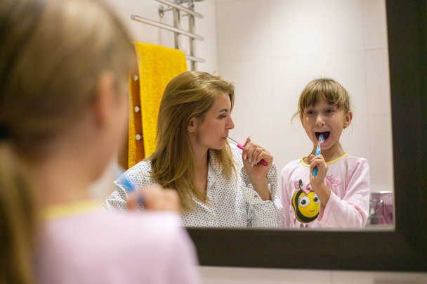 blonde little girl with a young beautiful mother cleans teeth in front of a bathroom mirror. - Valokuva, kuva