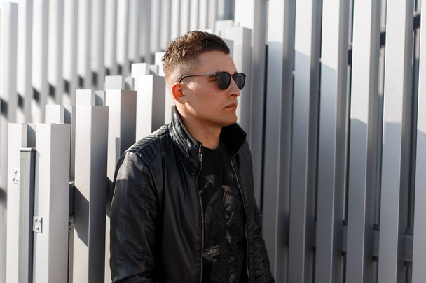 Fashionable hipster man with sunglasses in a stylish black leather jacket with a pullover stands near the metal wall - 写真・画像
