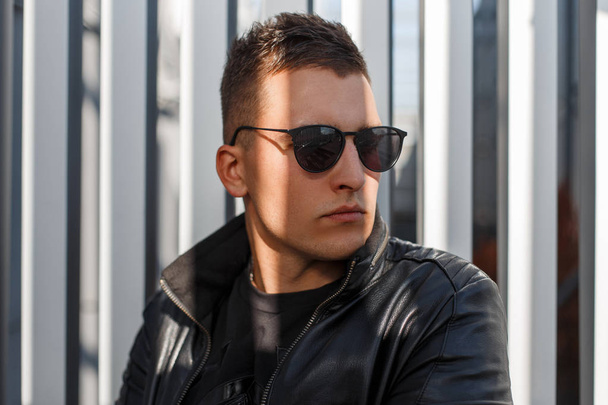 Portrait of an attractive stylish young hipster guy in dark sunglasses and a black leather jacket against a gray metal wall on a sunny autumn day. Stylish man. Close-up  - Photo, Image