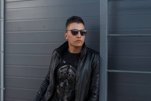 Handsome hipster young model man in black stylish sunglasses and black trendy clothes and fashion sweatshirt posing near a metal gray wall. Attractive urban fashionable guy - Photo, Image