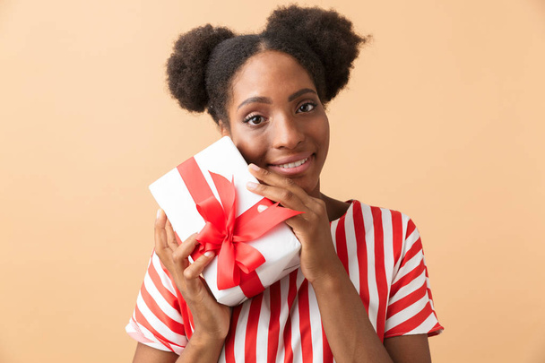 ベージュの背景の上分離された赤の弓とプレゼント ボックスを保持しているカジュアル衣料に愛らしいのアフリカ系アメリカ人女性 - 写真・画像