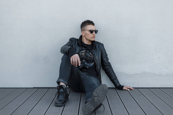 Knappe jonge hipster man in zwarte zonnebril, zwarte jas, stijlvolle pullover en zwarte jeans buitenshuis zittend op de houten vloer in de buurt van de grijze muur op een warme herfst dag. Mooie stijlvolle man - Foto, afbeelding