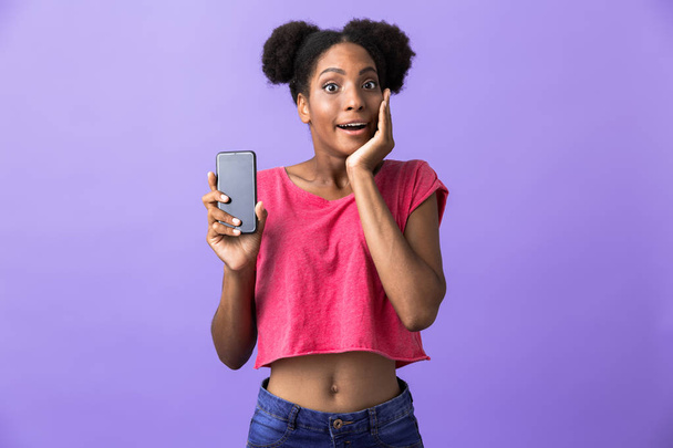 Photo of excited african american woman smiling and holding mobile phone isolated over violet background - Zdjęcie, obraz