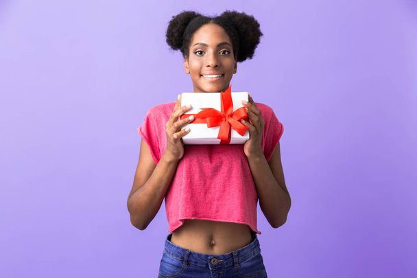 Foto pozitivní afroamerické ženy, usmívající se a držení dárkové krabičce s červenou mašlí izolované fialové pozadí - Fotografie, Obrázek
