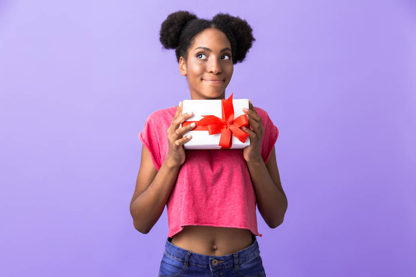 Photo of happy african american woman smiling and holding present box with red bow isolated over violet background - Valokuva, kuva