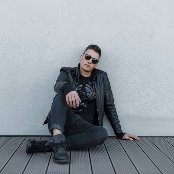 Attractive young hipster man in black sunglasses, black jacket, stylish sweatshirt and trendy jeans, rest and sits on a wooden floor outdoors near a gray wall. Fashionable modern guy - Foto, imagen