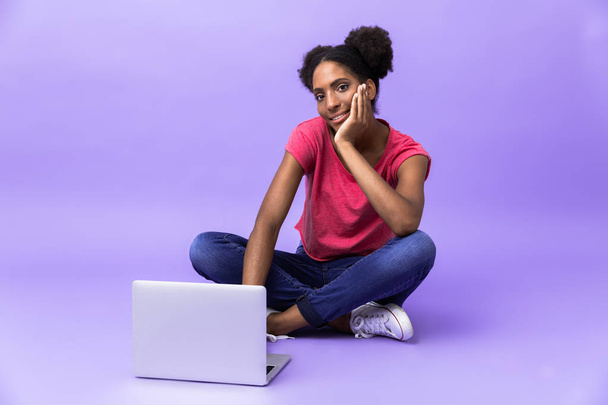 Photo of adorable african american woman smiling and using silver laptop, while sitting on floor with legs crossed isolated over violet background - Fotografie, Obrázek