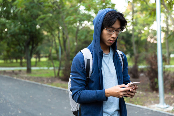 Image of young asian guy in casual wear and eyeglasses using smartphone while walking through green park - 写真・画像