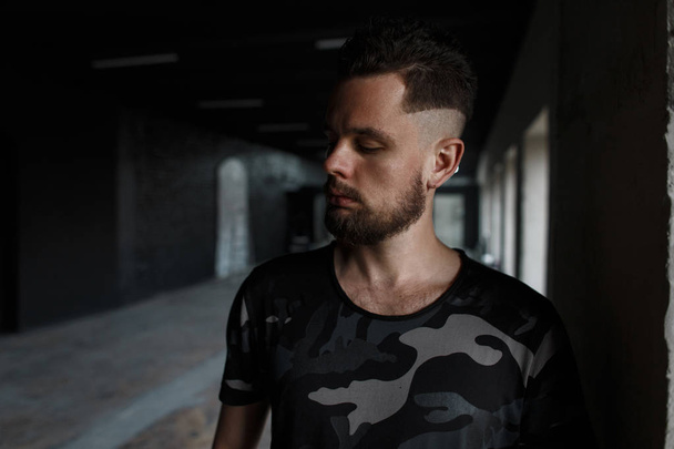 Handsome young man with a stylish hairstyle and brutal beard in a modern fashionable military t-shirt posing in a dark indoors. Attractive guy - 写真・画像