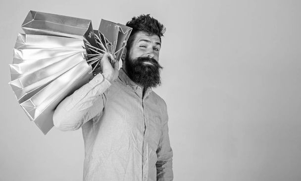 Hipster on winking face shopping. Shopping concept. Guy shopping on sales season, carries bunch of bags on shoulder. Man with beard and mustache holds shopping bags, grey background - Фото, изображение