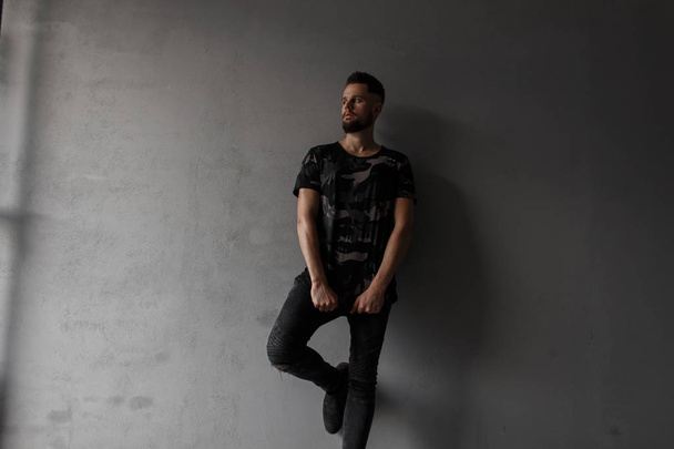 Αρκετά ελκυστικό νεαρό άνδρα με ένα μοντέρνο χτένισμα και βάναυση γένια σε ένα στρατιωτικό κομψό πουκάμισο, μαύρο τζιν και αθλητικά παπούτσια μόδας μαύρο, θέτοντας κοντά σε ένα γκρίζο τοίχο. Κομψό σύγχρονο άντρα - Φωτογραφία, εικόνα