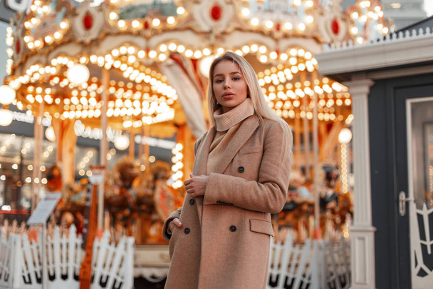 Piękna młoda moda Kobieta w stylowe jesienny płaszcz i vintage swetra w parku rozrywki na tle magiczny świąteczny jasne światła. Blond dziewczynka elegancki miejski moda. - Zdjęcie, obraz