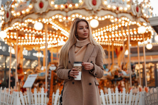 Hezká Mladá krásná žena v stylové podzimní kabát a vintage pletený svetr pití kávy v městském parku na pozadí magického jasná světla. Módní elegantní dívka blondýna - Fotografie, Obrázek