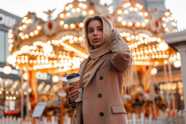 Young pretty woman in a coat and a vintage knitted sweater holds a cup of coffee and walks in an amusement park on the background of a carousel in the city. Elegant cute girl. - 写真・画像