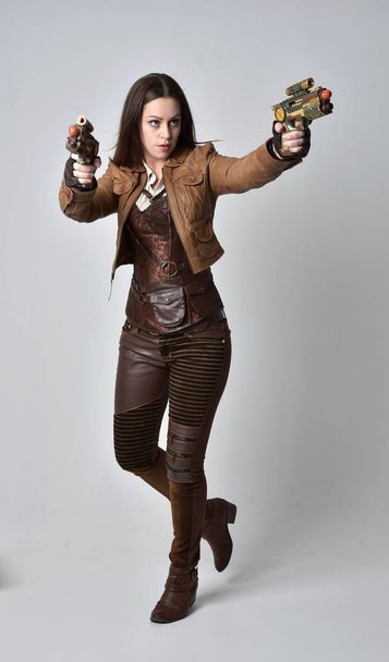 full length portrait of brunette  girl wearing brown leather steampunk outfit. standing pose holding a gun, on grey studio background. - Foto, Imagem