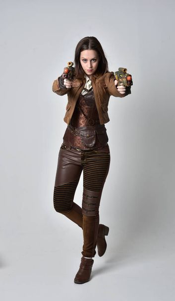 full length portrait of brunette  girl wearing brown leather steampunk outfit. standing pose holding a gun, on grey studio background. - Fotó, kép