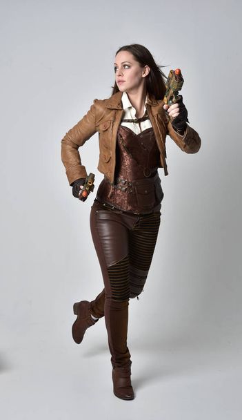 Ganzkörperporträt eines brünetten Mädchens im braunen Leder-Steampunk-Outfit. Stehende Pose mit Pistole, auf grauem Studiohintergrund. - Foto, Bild