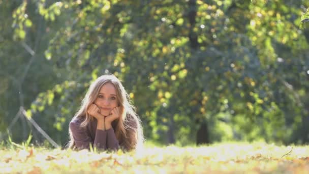 Дівчина в парку. Весела дівчина лежить на траві і обережно дивиться в камеру. Вона мріє, її посмішка мила і любляча. Її руки на щоках. На задньому плані дерева
. - Кадри, відео