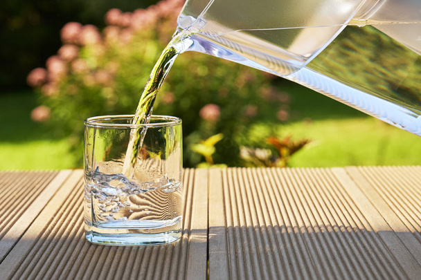 Wylewanie jasne filtrowanej wody z dzbanem filtracji do kieliszka na tle ogród zielony lato w ciepłej słoneczny letni dzień - Zdjęcie, obraz