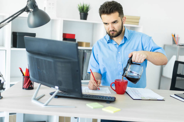 Een jonge man staat in het kantoor aan de computer tafel en brouwt zijn koffie. - Foto, afbeelding