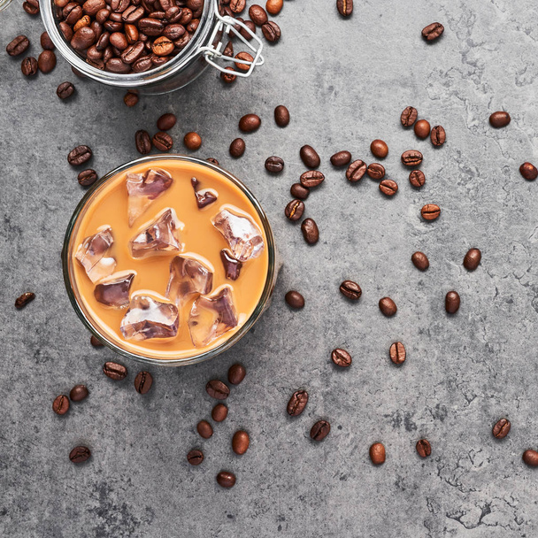 Chocolate, vanilla, caramel or cinnamon iced coffee in tall glass. Cold brewed iced coffee in glass and coffee beans in glass jar on grey concrete background. Top view, square crop. - Photo, image