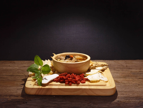 Schüssel mit chinesischer Suppe und pflanzlichen Medikamenten auf Holz gegen Schwarz - Foto, Bild