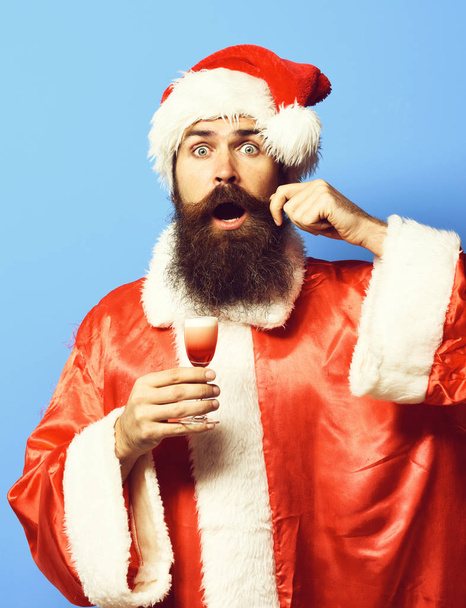 handsome bearded santa claus man with long beard on surprised face holding glass of alcoholic shot in christmas or xmas sweater and new year hat and touching mustache on blue studio background - Фото, изображение