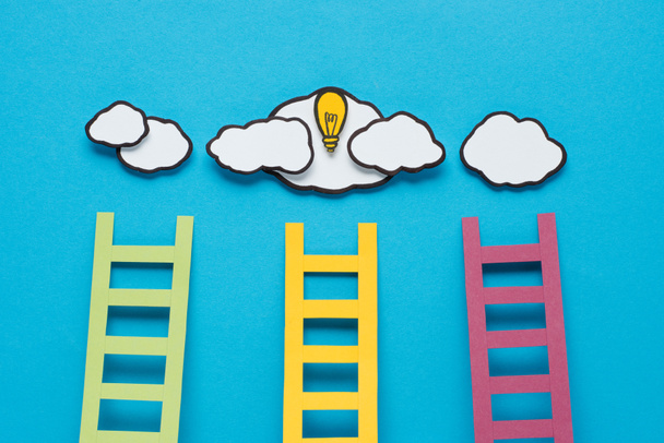 bovenaanzicht van kartonnen ladders met gloeilamp en wolken op blauwe achtergrond, ideeën concept - Foto, afbeelding