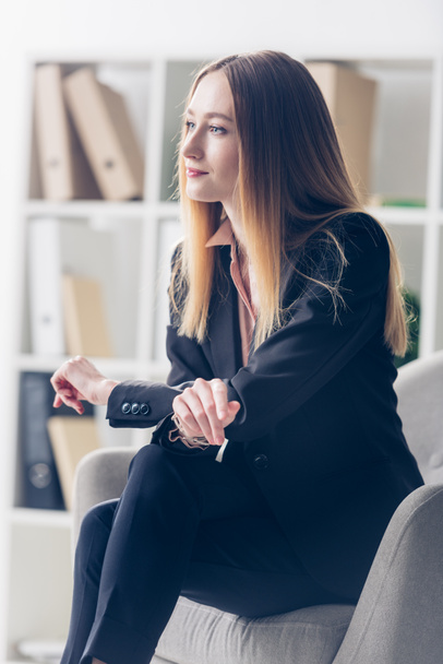 side view of attractive businesswoman in suit sitting on armchair and looking away in office - Foto, imagen