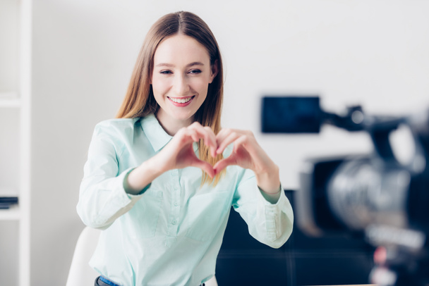 happy attractive female video blogger recording vlog and showing heart with fingers in office - Fotografie, Obrázek