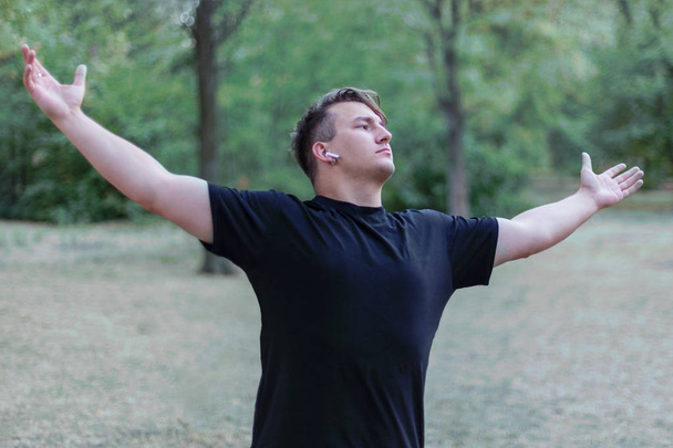 Young handsome caucasian man widely stretches out his hands (arms), at the park background. Wears casual (black t-shirt), white earphones, earring, ring. Stylish and attractive one. Outdoor, copy space. - Foto, Bild
