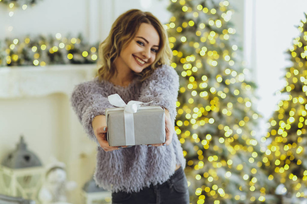 Young beautiful blonde in a gray sweater with a gift on hands near the New Year, Christmas tree with gifts - Zdjęcie, obraz