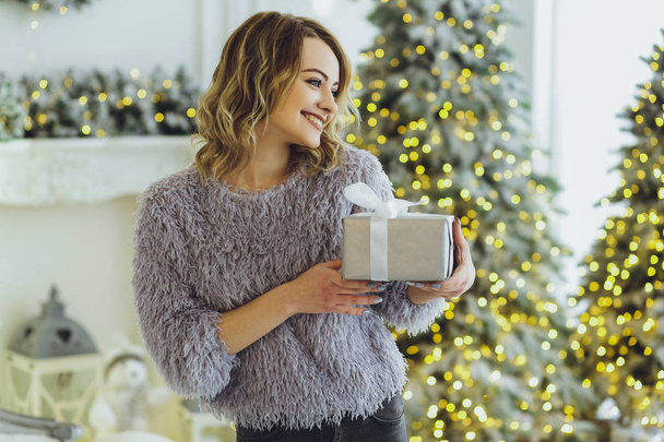Young beautiful blonde in a gray sweater with a gift on hands near the New Year, Christmas tree with gifts - Foto, Imagem