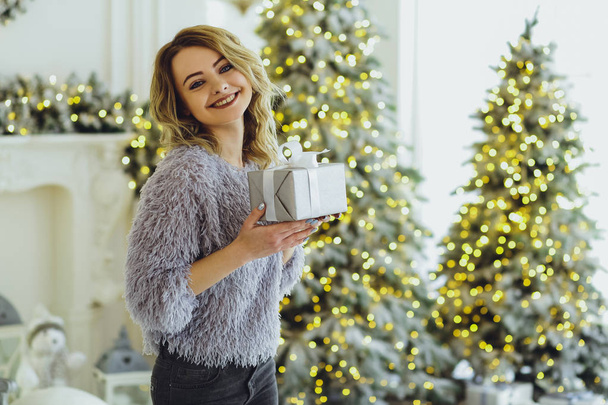 Young beautiful blonde in a gray sweater with a gift on hands near the New Year, Christmas tree with gifts - Foto, Imagen