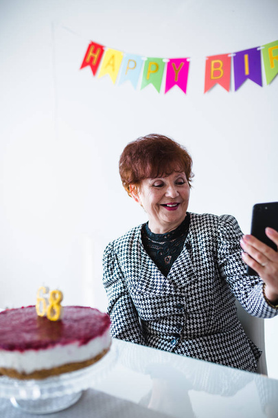 Mulher sênior fazendo selfie perto da mesa com bolo enquanto celebra o aniversário em casa
 - Foto, Imagem
