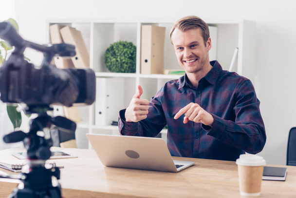 smiling handsome male video blogger recording vlog and showing thumb up in office - Foto, Bild