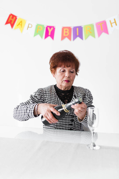 Senior woman opening bottle of champagne while celebrating her birthday at home with colorful fabric bunting on white wall background - Photo, image
