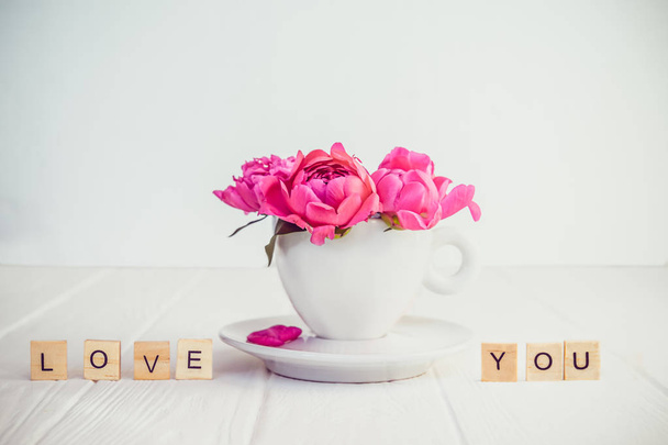 Close up light pink peony flowers bouquet in a decorative cup and saucer and message I love you spelled in wooden blocks on white wooden background. Valentines day, good morning concept. Copy space - Φωτογραφία, εικόνα
