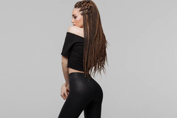 Beautiful young woman with dreadlocks keeping hand on waist and looking at camera while standing on light gray background - Photo, Image
