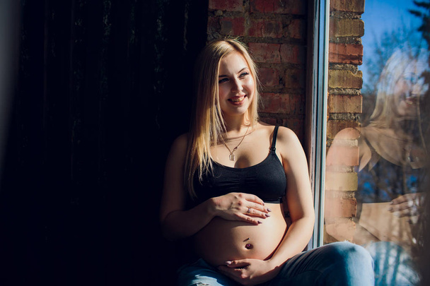 Young beautiful pregnant woman sitting on windowsill at home - Фото, изображение