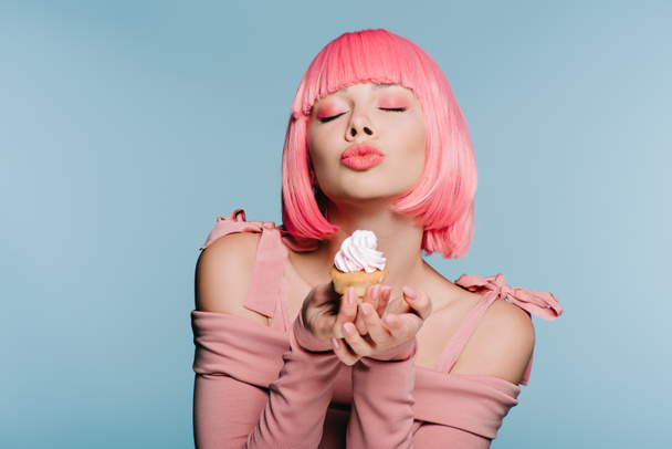 stylish girl in pink wig holding sweet cupcake and blowing kiss isolated on blue - Foto, afbeelding
