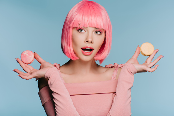 attraktive schockierte Mädchen in rosa Perücke posiert mit Macarons isoliert auf blau - Foto, Bild
