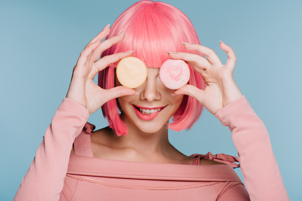 atractiva chica sonriente en peluca rosa posando con dos macarrones aislados en azul
 - Foto, Imagen