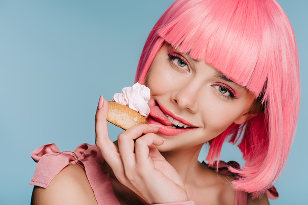 beautiful happy girl in pink wig licking sweet cupcake isolated on blue - Zdjęcie, obraz