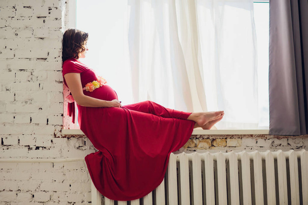 Pregnant woman in a red dress lying on the window female - Fotó, kép