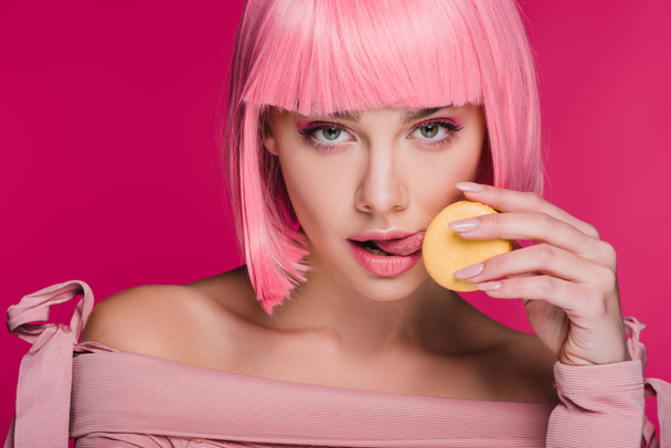 attractive girl in pink wig licking yellow macaron isolated on pink - Photo, Image