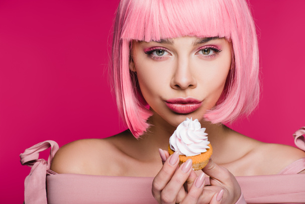 Atrakcyjna dziewczyna w różowych peruk trzymając pyszne ciastko z buttercream na różowym tle - Zdjęcie, obraz