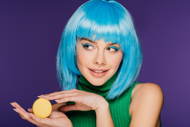 χαμογελώντας νεαρή γυναίκα σε μπλε περούκα ποζάρει με κίτρινο macaron απομονώνονται σε μωβ - Φωτογραφία, εικόνα